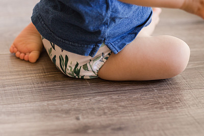 一个一岁大的男孩在地板上玩耍，学习走路，只穿着叶子图案的布尿布和蓝色衬衫