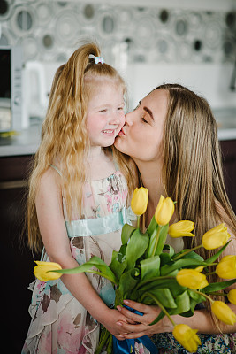 女儿在家里的厨房里向妈妈祝贺，并送上一束黄色的郁金香花。母亲节的概念。妈妈和小女孩拥抱亲吻。妇女节贺卡。特写镜头。