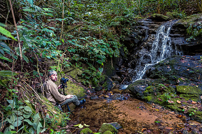 男子拿着三脚架和相机坐在瀑布前拍照。男摄影师在森林里拍摄美丽的瀑布。