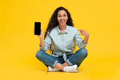 快乐的年轻女性指着智能手机的设计模型，在黄色背景上宣传新的应用程序或网站