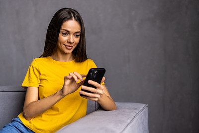 年轻漂亮的女人坐在客厅的沙发上，使用现代智能手机，在家打电话或订购。