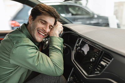 快乐的年轻人坐在驾驶座上，抱着他的新车方向盘，闭上眼睛微笑，复制空间