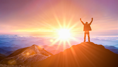 当太阳开始落山时，一名男子站在山顶。成功的商业领导。目标、希望和抱负的概念。日出山背景上的男性剪影