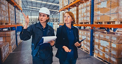 年轻的白人女性仓库工程师工人，成熟的成年女性经理在工厂仓库走动和讨论谈话。物流行业业务，行业工作，或人在工作的概念