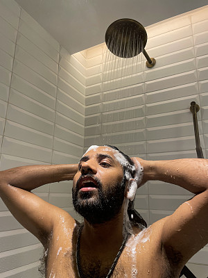 印度男子在淋浴间洗头，在流水下冲洗头发上的肥皂泡沫，白色斜边墙砖呈水平堆叠图案，重点放在前景
