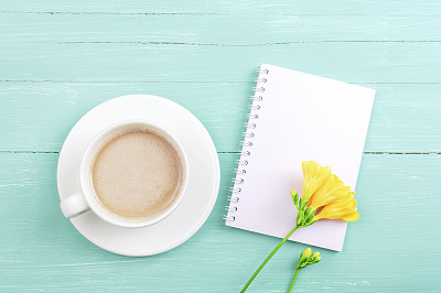 空白记事本和咖啡杯在绿松石木制桌子与小苍兰黄色的花。俯视图，平躺，模型