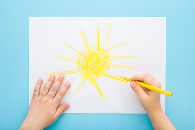 小孩子手拿黄色铅笔，在淡蓝色的桌子背景上白纸上画着大大的太阳。柔和的颜色。特写镜头。视角镜头。孩子创造时间。自顶向下视图。