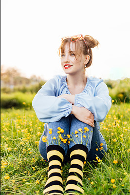 一名年轻女子穿着蓝色上衣、牛仔裤和黑黄相间的条纹袜子，袜子里插着鲜花，坐在盛开的草地上。动画风格。蜜蜂保护概念，开花季节。