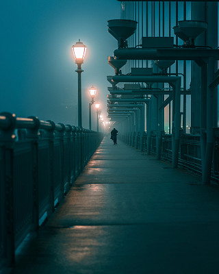 一名男子在雾蒙蒙的夜晚过桥，前景是灯柱