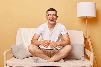 出望外的黑发成年男子穿着休闲服,坐在沙发上,靠着米色的墙,心情很好