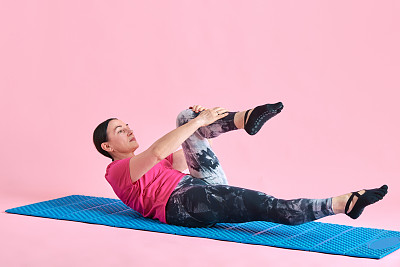 成熟美丽的女人躺在健身matt和伸展腿，膝盖在粉红色的工作室背景