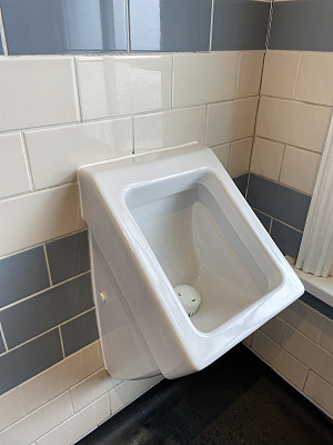公厕厕所的小便池特写，现代方形设计的白色陶瓷小便池靠近窗户，干净卫生的小便池安装在交错的砖粘结墙砖和混凝土地板上，重点放在前景