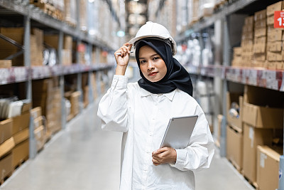 Portrait of Asian muslim female headscarf hijab wear hard hat holding digital tablet checklist product shelves warehouse. woman worker inspecting goods industry. Logistic concept.