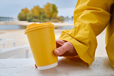 女人手里拿着咖啡的黄色纸杯。是时候在城市里喝咖啡了。咖啡外带。享受这一刻，休息一下。一次性纸杯特写。美味的热饮。空白的文字，模型