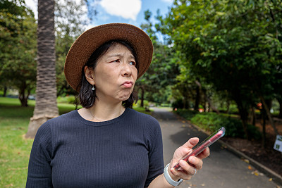 一个亚洲女人在公园里拿着手机，一脸困惑