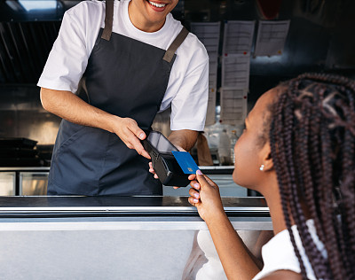 一名快餐车老板拿着POS机，而一名女顾客用非接触式卡支付