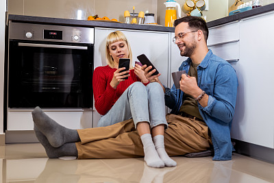 一对幸福的夫妇坐在厨房的地板上看着他们的手机，微笑着