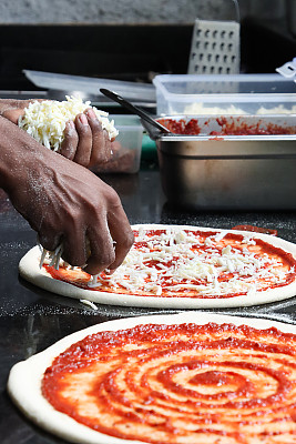 面面俱到的Pizzaiolo(披萨厨师)将磨碎的马苏里拉奶酪洒在未煮熟的披萨底座上，玛格丽塔披萨富含番茄马里纳拉酱，磨碎的马苏里拉奶酪，商业厨房设置，重点放在前景