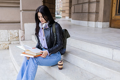 年轻漂亮的女大学生背着双肩包坐在大学楼梯上。她戴着耳机，穿着皮夹克，等待着大学课程的开始。