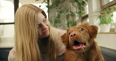 一位女士和心爱的宠物狗新斯科舍省鸭鸣猎犬一起躺在沙发上，愉快地抚摸着它