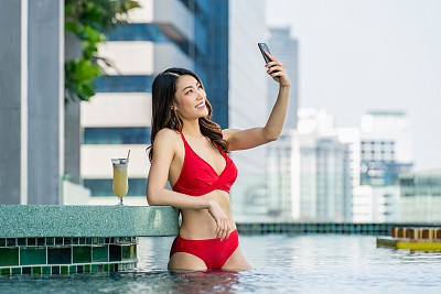 城市风光自拍:女子开心地笑着，在屋顶泳池边用手机拍下这一刻，旁边有饮料，背景是城市风光。