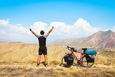 近距离后景，鼓舞人心，兴奋，快乐的高加索男性骑着红色的旅游自行车在荒凉的山区，举手自信，无忧无虑的生活方式