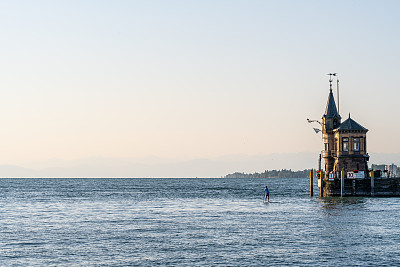游客站在站立式桨板上经过灯塔和海港入口，入口处挂着康斯坦茨旗。康斯坦茨，康斯坦茨湖，巴登-符腾堡州，德国，欧洲。
