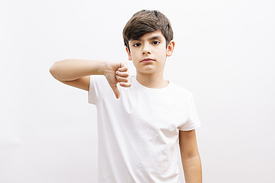 穿着白色t恤的小男孩站在白色孤立的背景上，看起来不高兴和愤怒，用拇指向下的手势表示拒绝和否定。糟糕的表达。