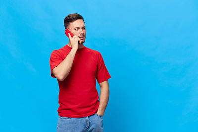 愤怒、恼怒、困惑、棕褐色、穿着红色t恤的英俊男子从电话中得到糟糕的财务报告，在蓝色背景下单独摆姿势。复制空间横幅模型。人们的生活方式理念