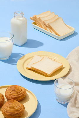 早餐概念，几种面包陈列在陶瓷盘子上，杯子和一瓶牛奶。蓝色的表面。美味健康食品的理念