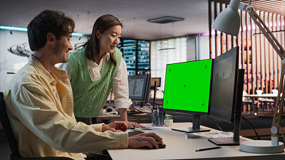 在游戏开发工作室办公室，白人男性游戏设计师使用绿屏台式电脑与亚洲女性项目经理交谈。不同的同事讨论新的RPG游戏