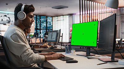 富有创造力的印度人的肖像坐在桌子上使用桌面电脑与模拟绿屏Chromakey。在游戏设计初创公司工作的男性关卡设计师，创造沉浸式游戏玩法