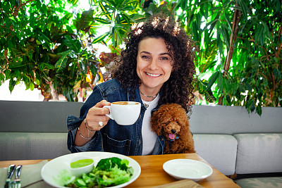 美丽卷曲的黑发女人的肖像，带着宠物在素食餐厅享受植物性蔬菜餐，环保的室内装饰着郁郁葱葱的绿色植物