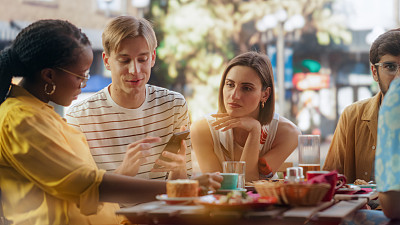 一位年轻的时尚男士与多民族朋友坐在露台上，用智能手机展示度假照片和社交网络信息。年轻的朋友们在咖啡馆里愉快地聊天