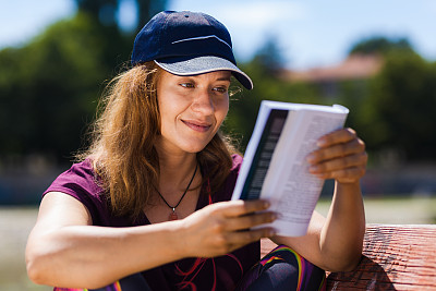在阳光明媚的公园里，一位戴着帽子、面带微笑的女子正在看书，在晴朗的天气里表现出轻松和满足。