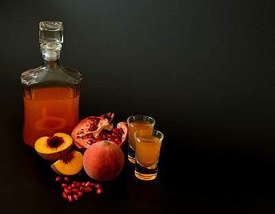 一瓶和两杯自制的水果利口酒，背景是黑色的，旁边是熟桃子和碎石榴。