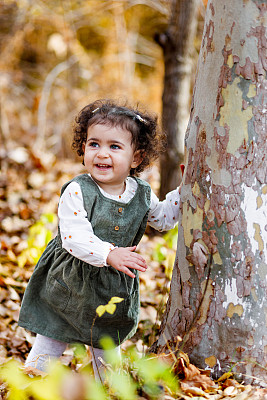 微笑的小女孩站在一棵树旁，抚摸着树皮，脚下的树叶