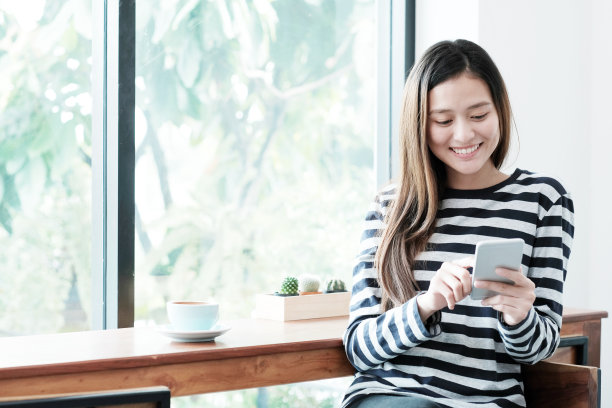 年轻的亚洲女子使用智能手机，而坐在窗口咖啡馆背景，人的技术和生活方式