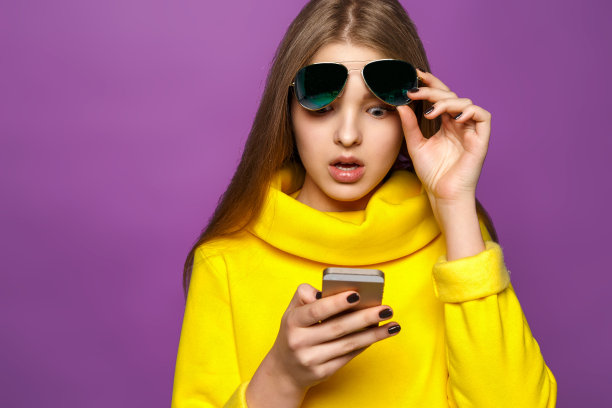 肖像惊讶的年轻女孩从智能手机短信在明亮的黄色毛衣，孤立在紫罗兰的背景