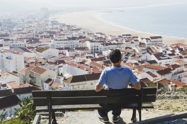 一个成熟的男人坐在长椅上，看着葡萄牙的纳扎尔小镇和海滩