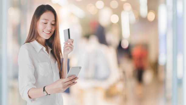 年轻美丽的亚洲女人用信用卡购物。在模糊的购物中心背景下拿着智能手机微笑。人们使用技术的概念。