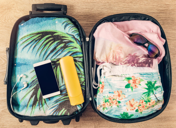 五颜六色的海滩配件与棕榈树的图片上的衣服和毛巾在旅行袋放松在炎热，异国情调的地方。木质地板背景上打开的手提箱。准备旅行的概念。