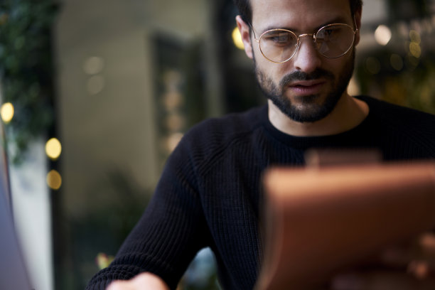 裁剪图像年轻的经济学家男性，戴着时尚的眼镜，一边在咖啡馆工作，一边在笔记本上读笔记。Ð“一个坐在咖啡店里，把想法写在现代笔记本上的大胡子嬉皮士。