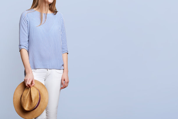 裁剪镜头的不认识的女性，条纹衬衫和白色裤子，持有草帽，穿着时尚的衣服，孤立在蓝色背景与复制空间为您的广告。