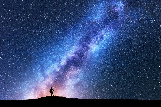 夜晚，在令人惊叹的银河中，拄着拐杖的人的剪影。空间背景。山峦上有人，银河灿烂，天空有星星。美丽的星系。旅行。星空