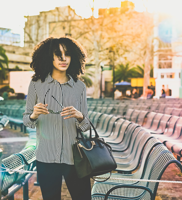 Beautiful,early,20’s,young,woman,of,mixed,Hispanic,and,African,American,ethnicity.,She,is,stunning,bouncy,natural,hair,and,is,happy,and,full,of,vitality.,She,is,in,a,city,environment,at,dusk,and,the,sunflare,filters,through.,She,is,a,confident,and,happy,millennial,with,a,infectiously,positive,attitude