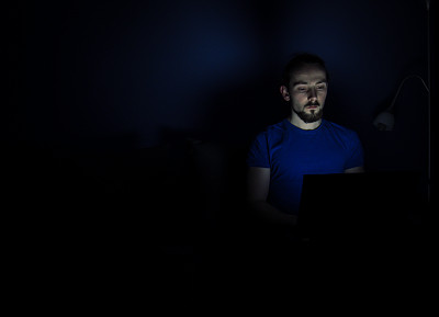 年轻人坐在床上，在黑暗中看着笔记本电脑的屏幕。