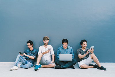 多民族的四名男子使用智能手机，笔记本电脑，数字平板电脑以及复制空间在蓝色的墙上。生活方式与信息技术小工具，教育，或社会网络的概念