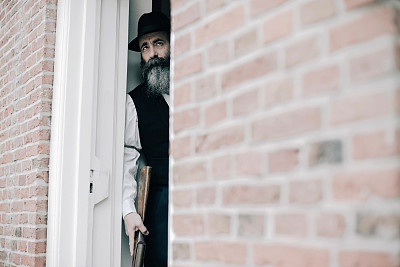 一个戴着帽子留着长胡子的神秘男子拿着步枪站在开着的门口。