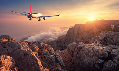 飞机飞行。风景与白色的客机，岩石，山脉，大海和橙色的天空与云彩日落。夏天的旅程。客机正在降落。商务旅行。商用飞机